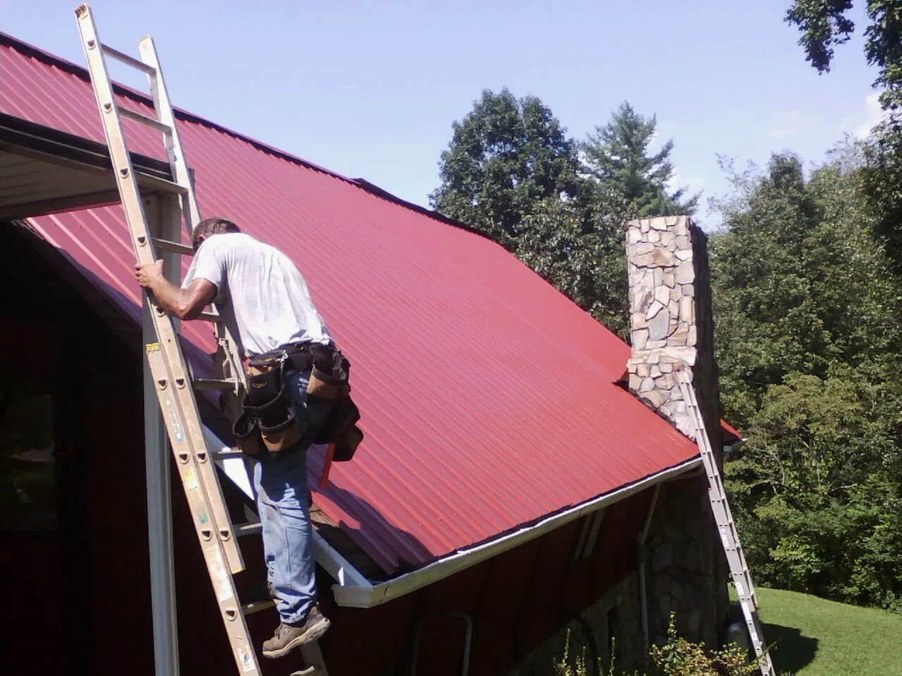 Roofer on a ladder beside a red metal roof and stone chimney — Soffit & Fascia Repair by Roofing Brothers in Leslie